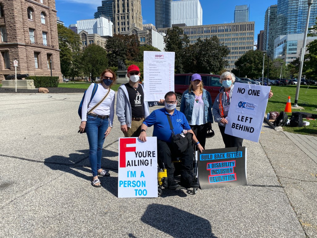 Image Below is of the 2021 Rally4ODSP Organizing Committee all wearing masks:  Front row seated Anthony Frisina, Back standing are Sherry Caldwell, Kyle Vose, Andrea Hatala and Catherine Manson. We are holding up rally signs that read, NO ONE LEFT BEHIND with the ODC logo, The ODSP Action Coalition logo with the graded ODSP report card below. Another sign next to Anthony reads Build Back Better, A Disability Inclusion Revolution.  Last rally sign reads F your Failing I'm a person too. 